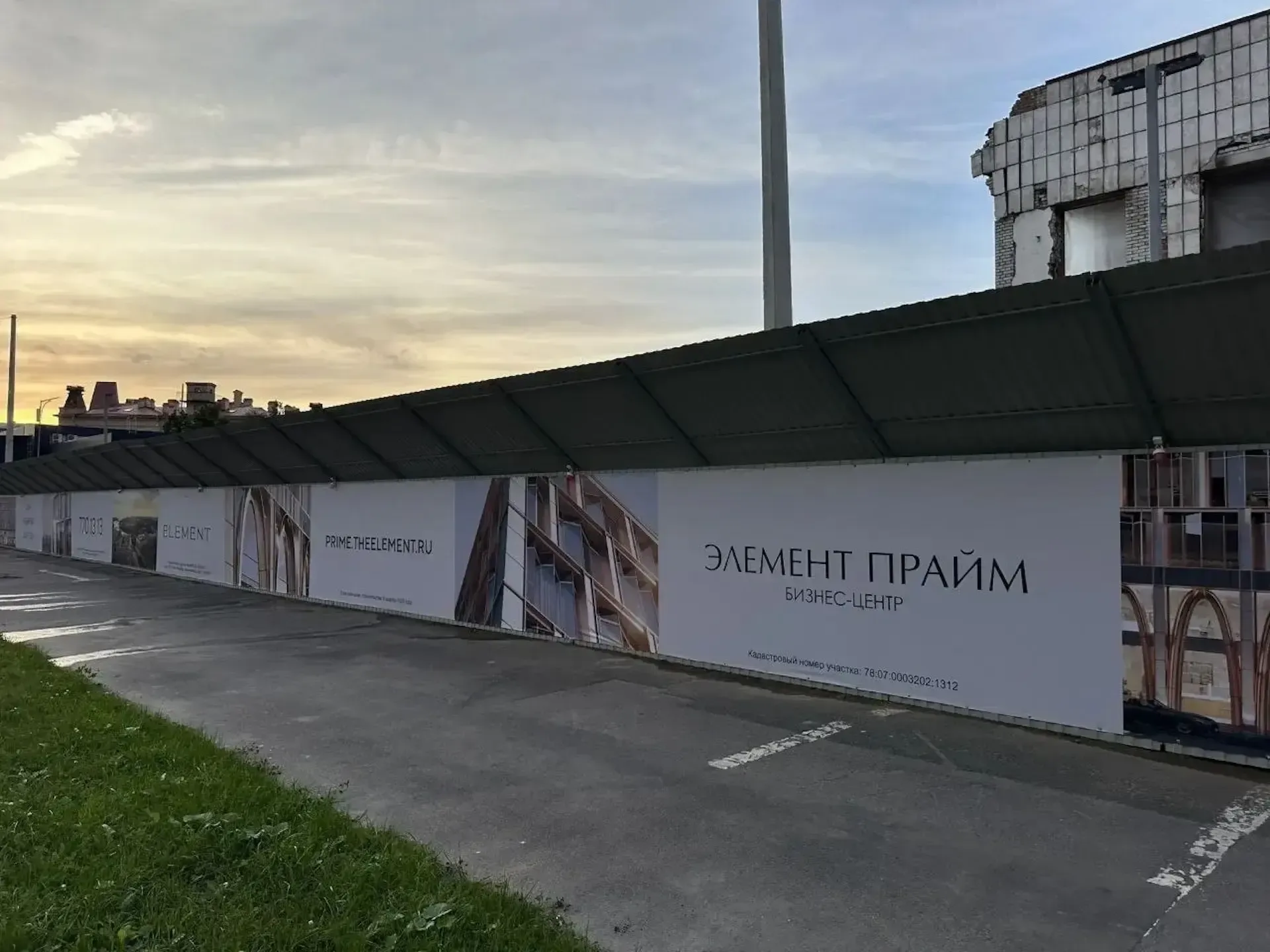Строительное ограждение вдоль проезда с надписью «Элемент Прайм бизнес-центр» и изображениями фасада, за ним здание со снятой обшивкой
