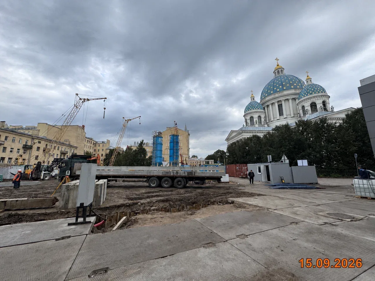 Стройплощадка «Фонтанки 130»:бетонные плиты временной дороги, техника и башенные краны, за деревьями синие купола Троицкого собора под пасмурным небом