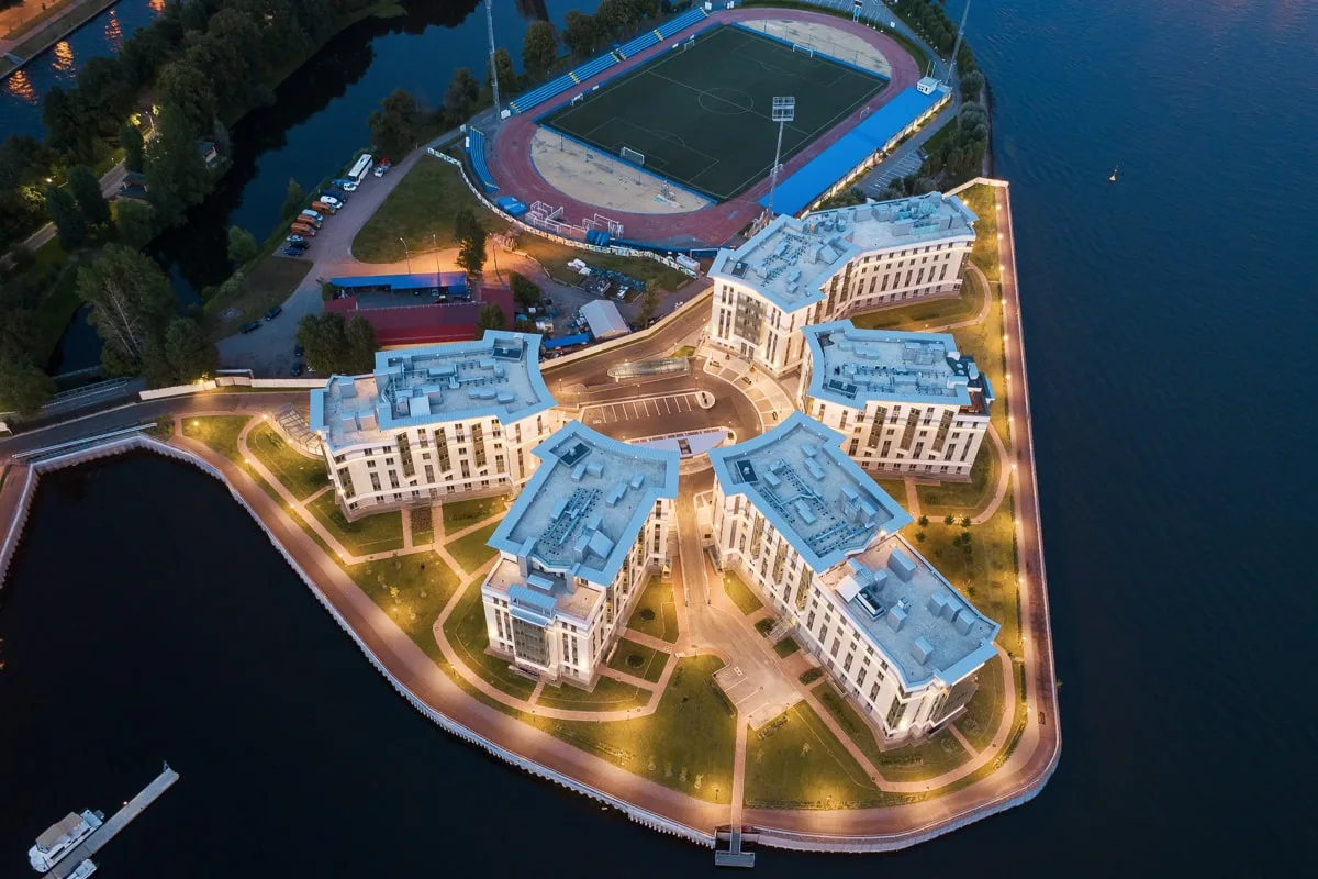 Royal Park строго сверху: корпуса расходятся веером от круглой площади, вокруг зелень двора, рядом чаша стадиона и вода